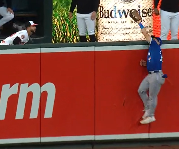 Daulton Varsho reaches well over the outfield wall to make a catch as an Orioles relief pitcher watches.