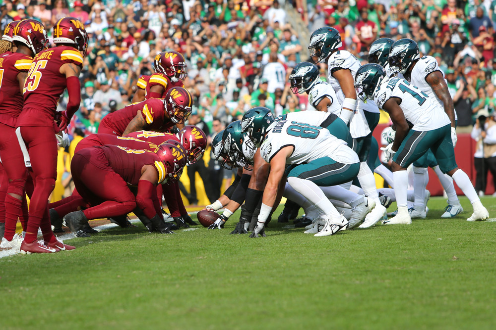 The Eagles, in white jerseys and green pants, prepare to execute their QB sneak "Tush Push" against a Commanders team wearing red uniforms.