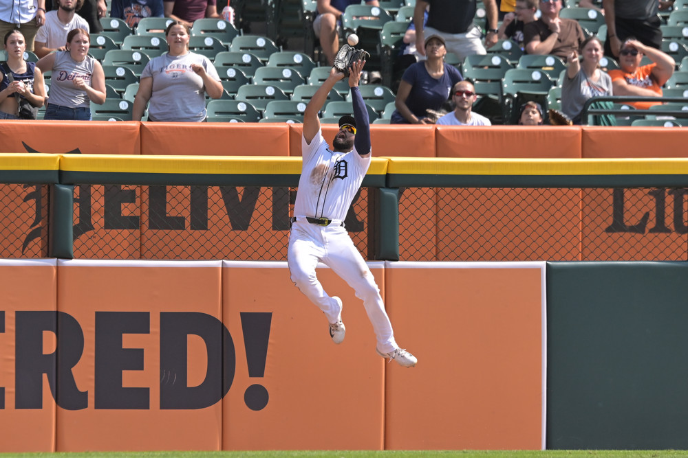 Riley Greene, in white jersey and white pants, leaps high at the outfield fence to make a catch and take a home run away from Ezequiel Tovar of the Rockies.