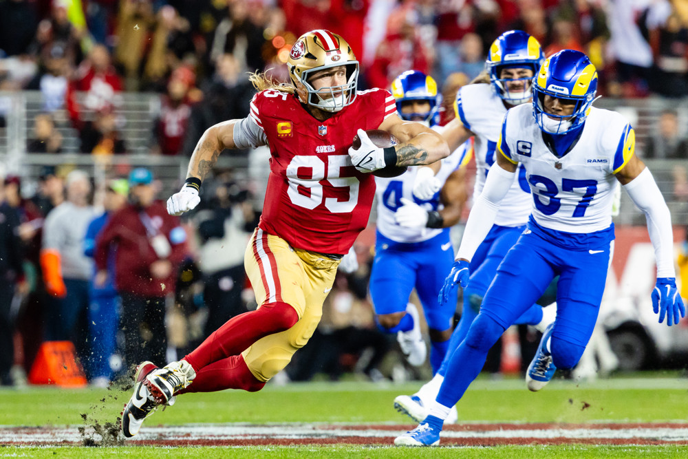 George Kittle makes a catch and runs upfield against the Rams. He's wearing a red jersey and white number 85, a gold helmet, gold pants and high red socks.