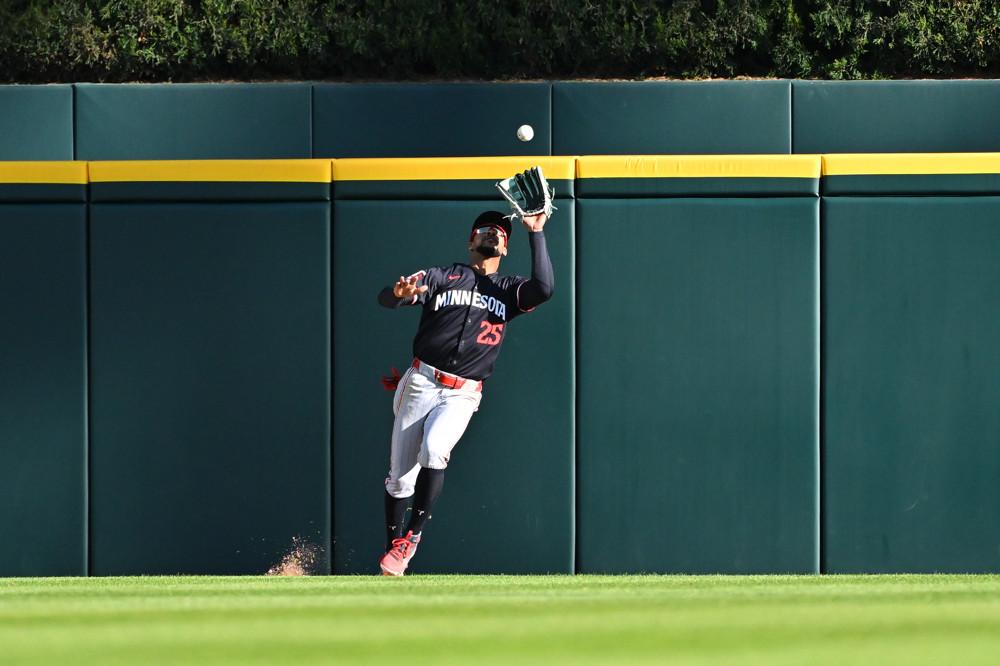 Byron Buxton is about to catch a fly ball. He's in a dark blue jersey with blue socks and white pants. He's holding his glove up with his left hand.