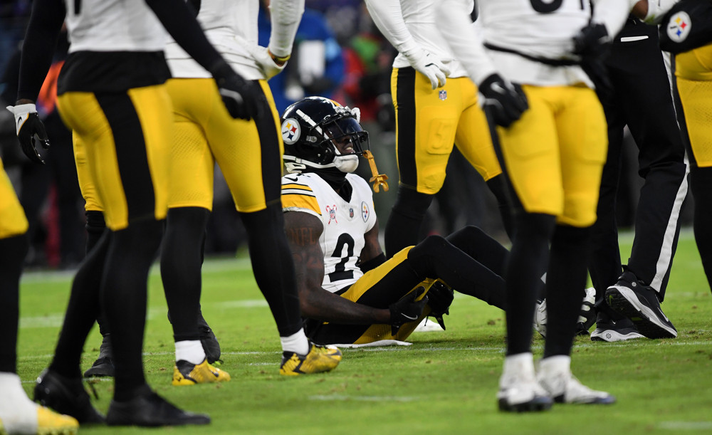 An injured member of the Pittsburgh Steelers sits on the turf while the rest of his team is standing