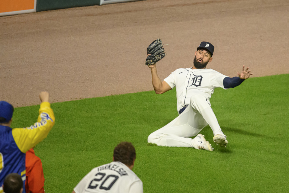 Riley Greene, in a white jersey with the Tigers logo and a black cap, makes a sliding catch with his glove up.