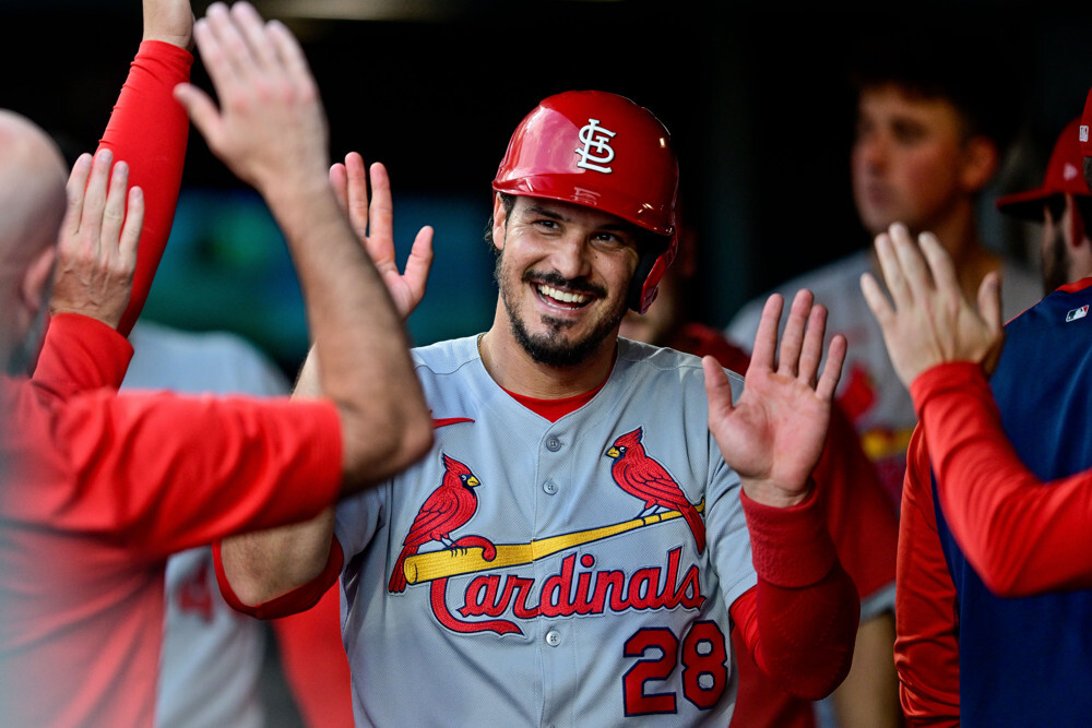 Nolan Arenado, wearing a Cardinals jersey and a red helmet with the STL logo, is giving high-5s to his teammates