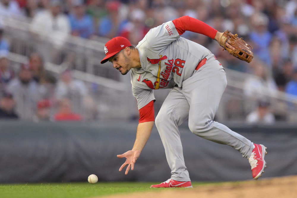 Nolan Arenado, in grey jersey with red lettering, a red cap, and a red undershirt, leans forward to field a baseball with his bare right hand