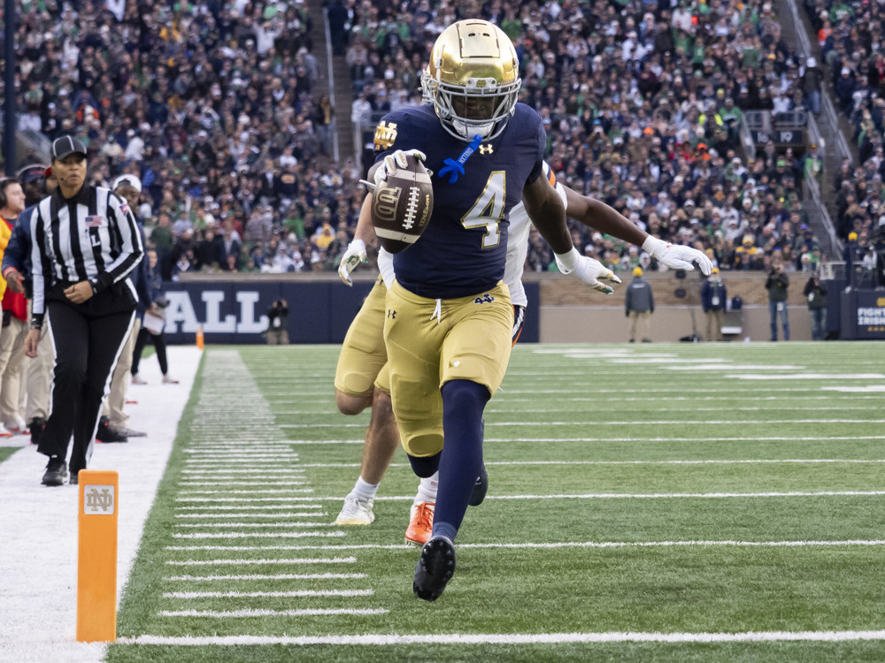 Jeremiyah Love, wearing a blue jersey and a gold helmet with a #4 on his jersey in white letters, holds the football out as he sprints and approaches the goal line for a touchdown