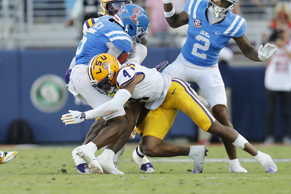 Mansoor Delane, wearing a yellow helmet, white jersey, and yellow pants, tackles a player around the waist, his helmet driving into the player's pants