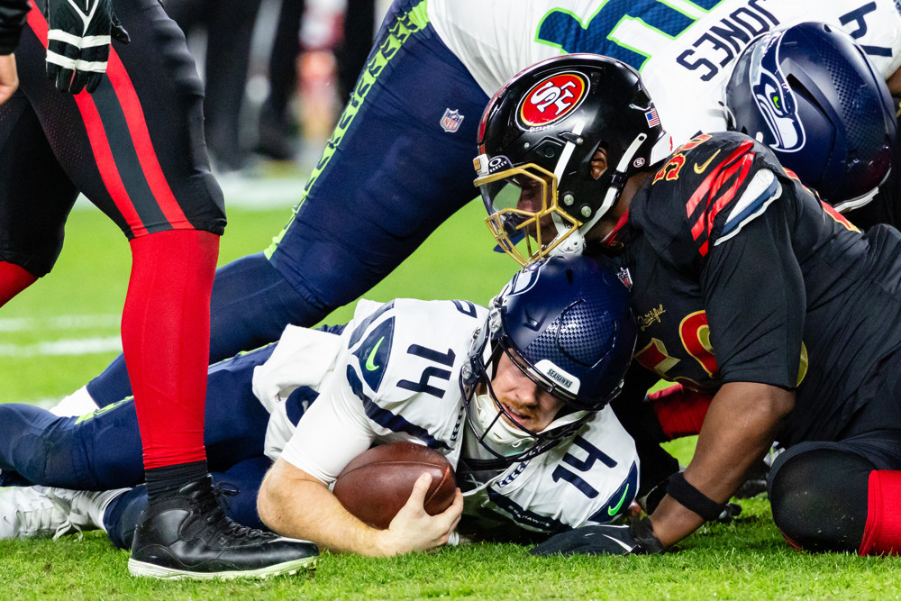 Sam Darnold, in white #14 jersey and blue helmet, protects the football while lying on his stomach as he's tackled by players on the 49ers