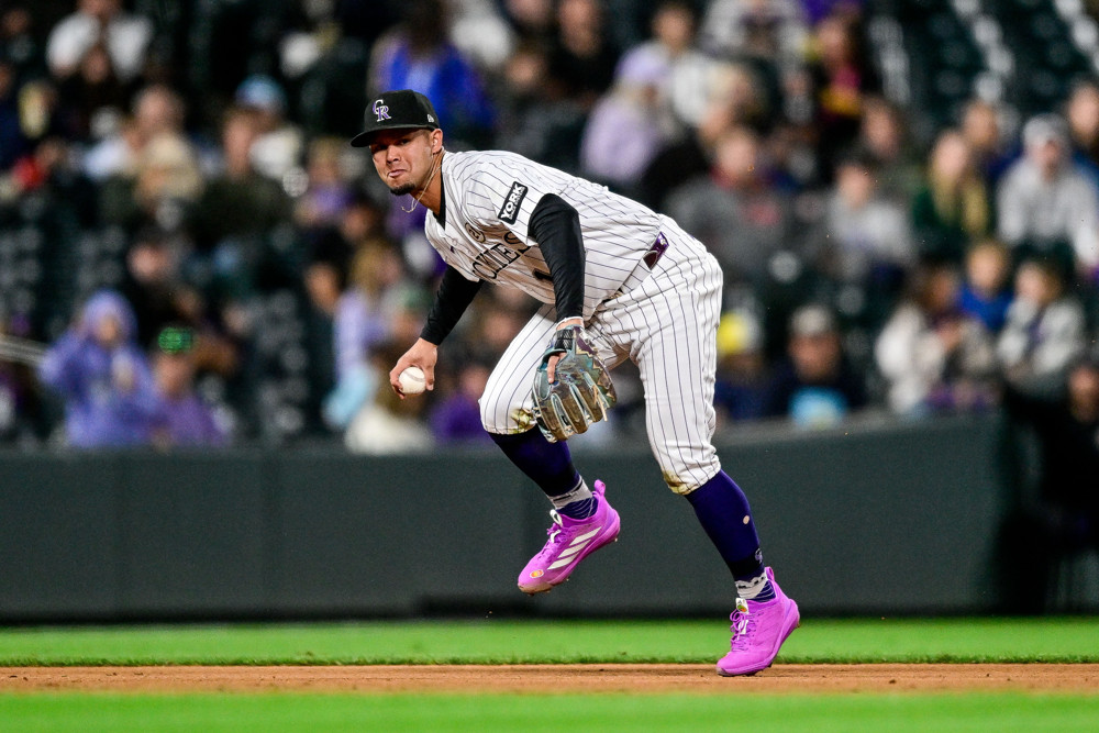 Ezequiel Tovar bends over to make a throw to first base, the ball held back in his right hand. He's wearing an all-white Rockies jersey with black socks and a black cap, with purple sneakers.