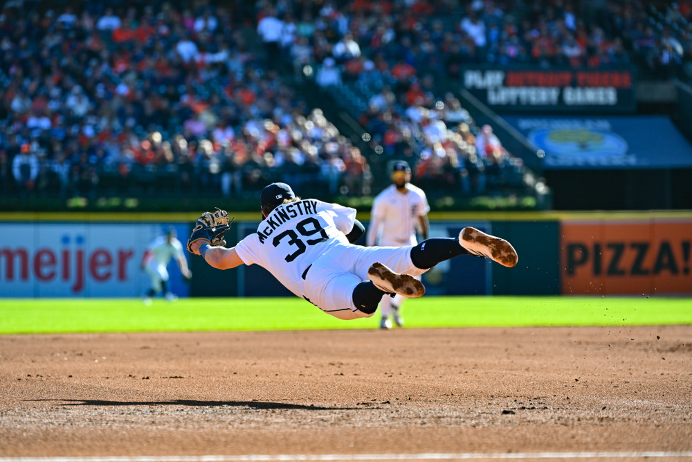 Zach Mckinstry, wearing a white Tigers jersey, dives with a full extension to his left to try to field a ball hit his way.