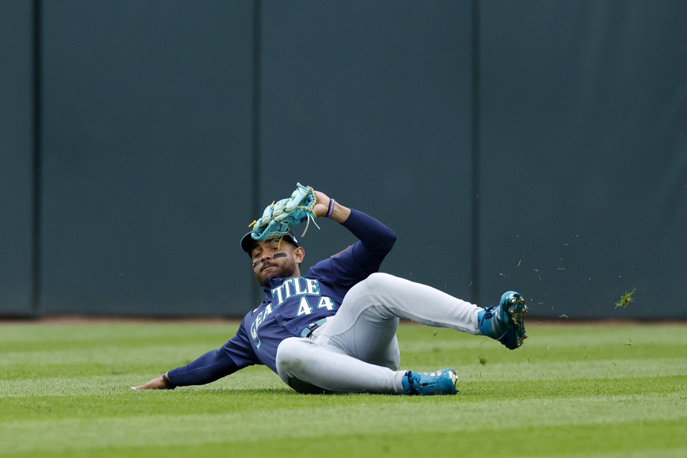 Julio Rodriguez, wearing a blue Mariners uniform and grey pants, slides forward with his glove on his left hand raised to try to make a catch.