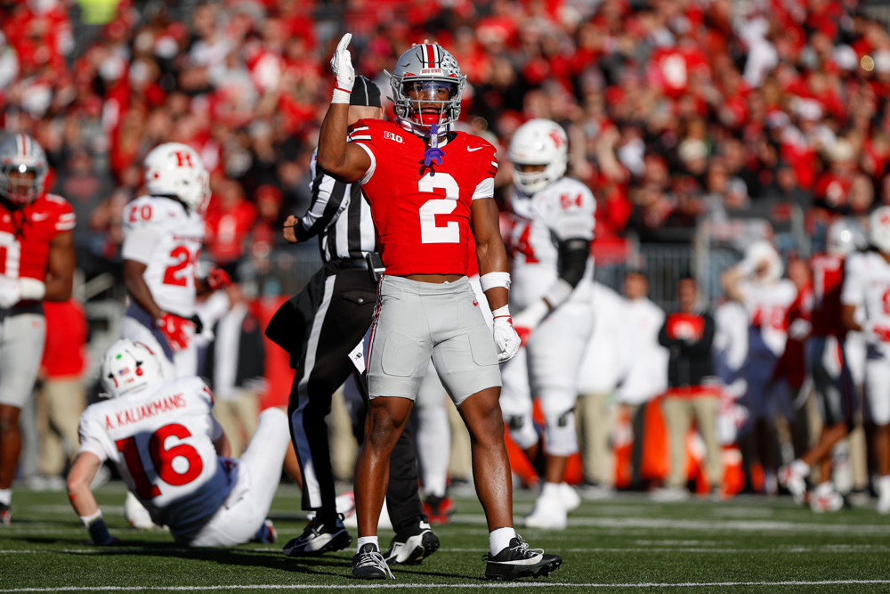 Caleb Downs, wearing a red Ohio State football jersey with a white #2, raises his arm as if he's about to make a chopping motion.