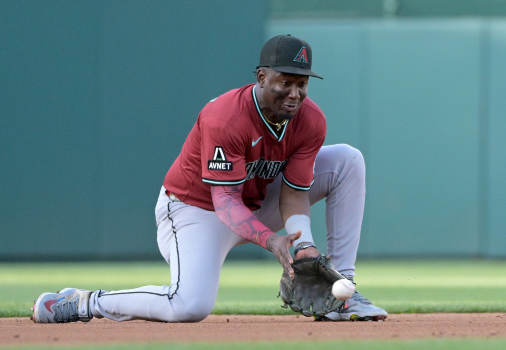 Geraldo Perdomo drops to one knee and props himself up on his right knee as he's about to field a ball. He's wearing a dark red baseball jersey and grey pants with a black Diamondbacks baseball cap.
