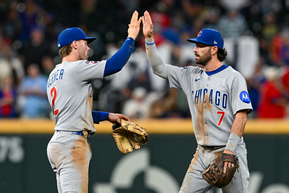 Nico Hoerner and Dansby Swanson give each other a high five. They are both wearing dirty grey Cubs uniforms and blue hats. Swanson has a beard. Both are high-fiving with their right hand.