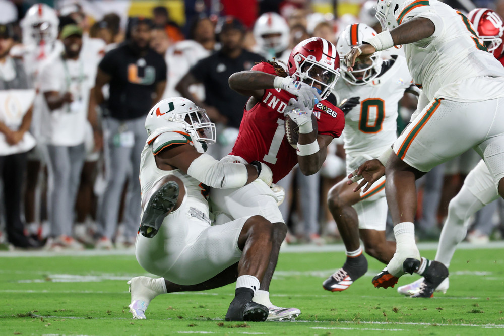 Rueben Bain Jr., from a crouched position and wearing an all white jersey with a Miami logo on his hemet, brings down a player wearing a #1 Texas Tech jersey who is struggling to stay upright.