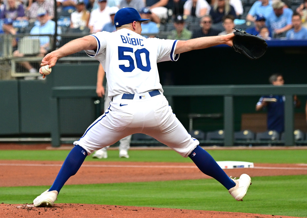 A pitcher, Kris Bubic, wearing a white jersey with 50 Bubic in blue, white pants and blue socks, reaches back with his left arm and steps forward with his right arm to throw a pitch.