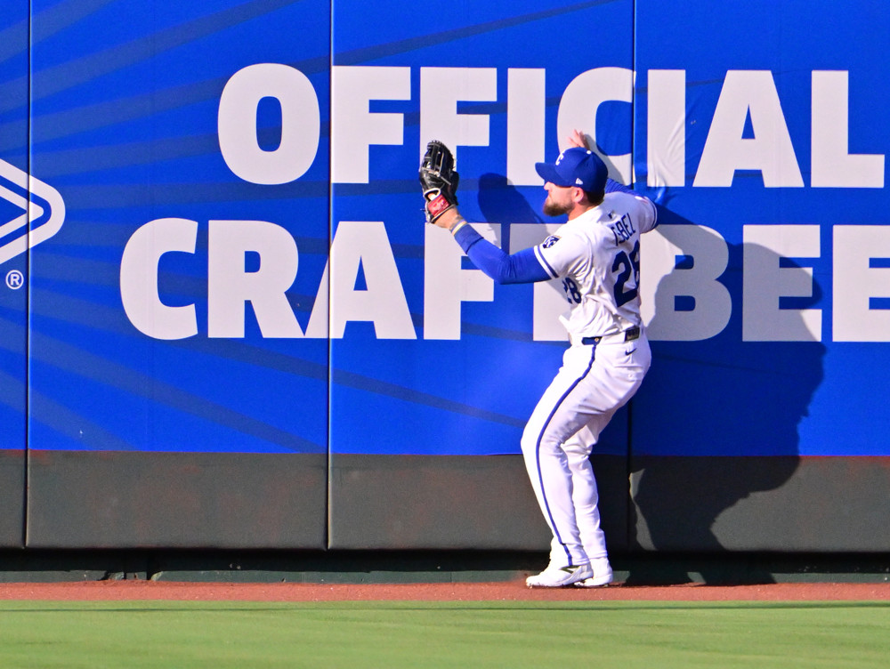 Kyle Isbel, wearing an all-white Royals uniform stands with his right side facing the fance, glove extended to make a catch in front of a sign advertising craft beer.