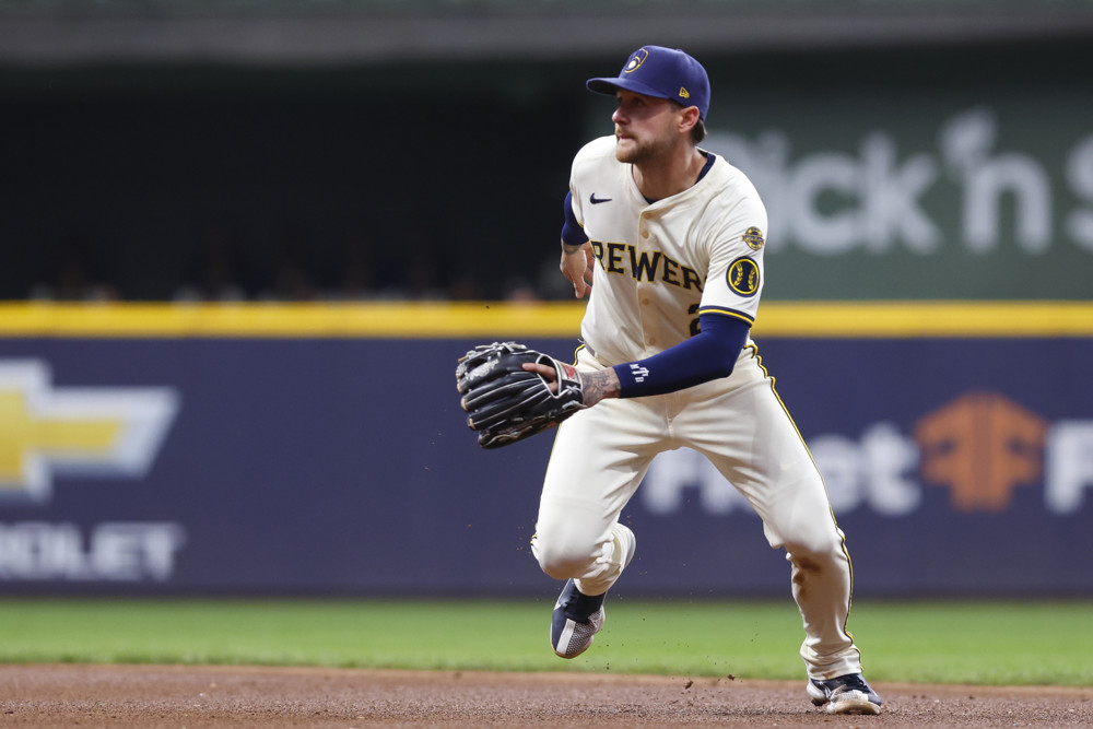 Brice Turang, in white Brewers jersey with Brewers in script across the front, and a blue cap, moves forward with his left arm in his glove, preparing to field a ball.