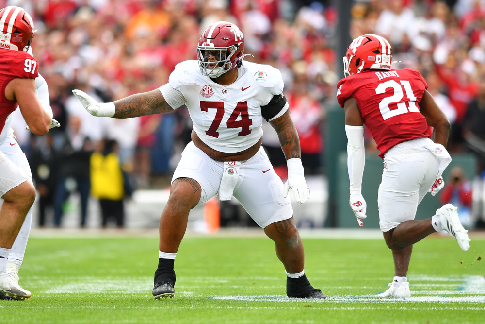 Kadyn Proctor, wearing a white #74 Alabama jersey and a red helmet, motions to his right with his right arm as he prepares for a snap.