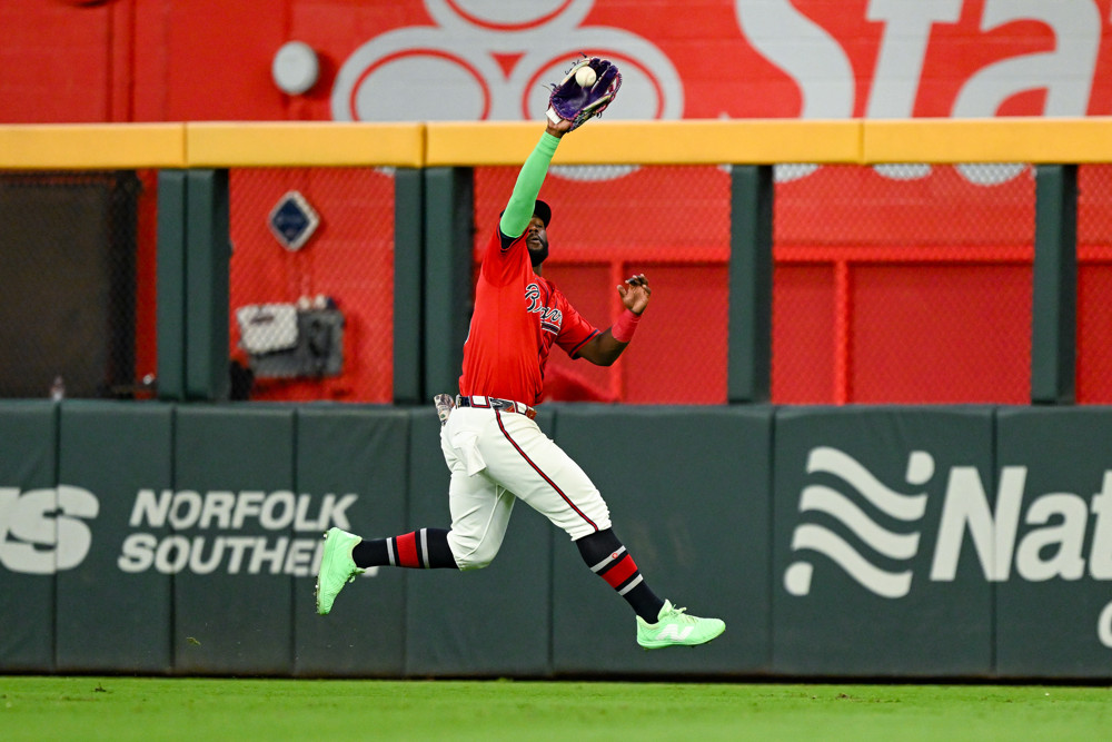 Michaerl Harris II, wearing a Red Braves jersey and white pants, with a green undershirt, reaches high in the air with his glove (on his right hand) to catch a fly ball as he retreats backwards.