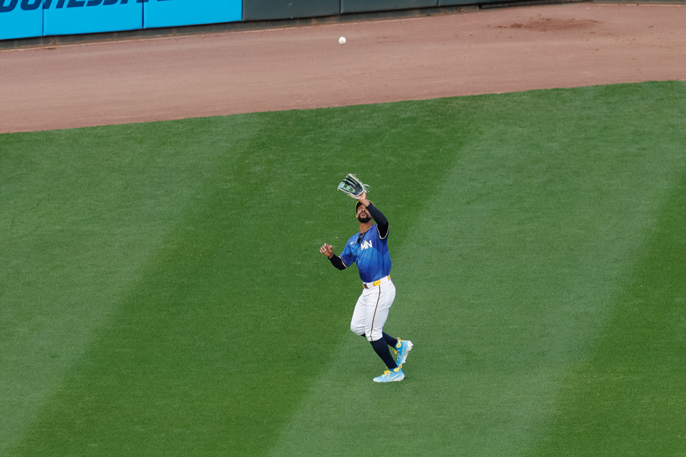 Byron Buxton, wearing a light blue uniform, white pants, and black socks, reaches up with his glove to make a catch in left center field.