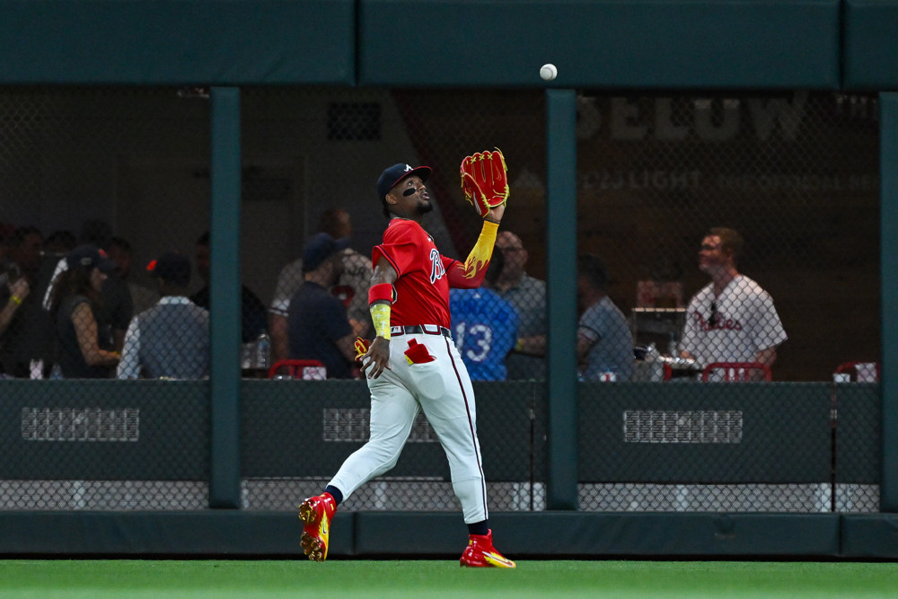 Ronald Acuna Jr. wearing a Red Braves jersey and white pants, retreats and reaches out with the glove on his left hand to catch a fly ball.