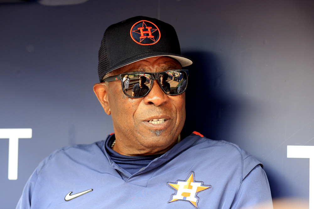 Dusty Baker, wearing a blue Astros pullover with an Astros logo, black sunglasses, and a black Astros cap, talks in the dugout.