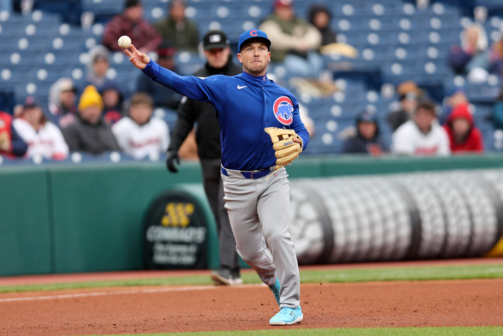 Alex Bregman, wearing a blue Cubs jersey, blue camp, and grey pants, makes a throw to first base from a 3/4 overhand position. The seats behind him are blue and mostly empty.